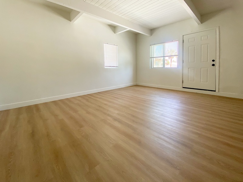 Empty living room with two windows, the front door and wood-look plank flooring under a beamed ceiling
